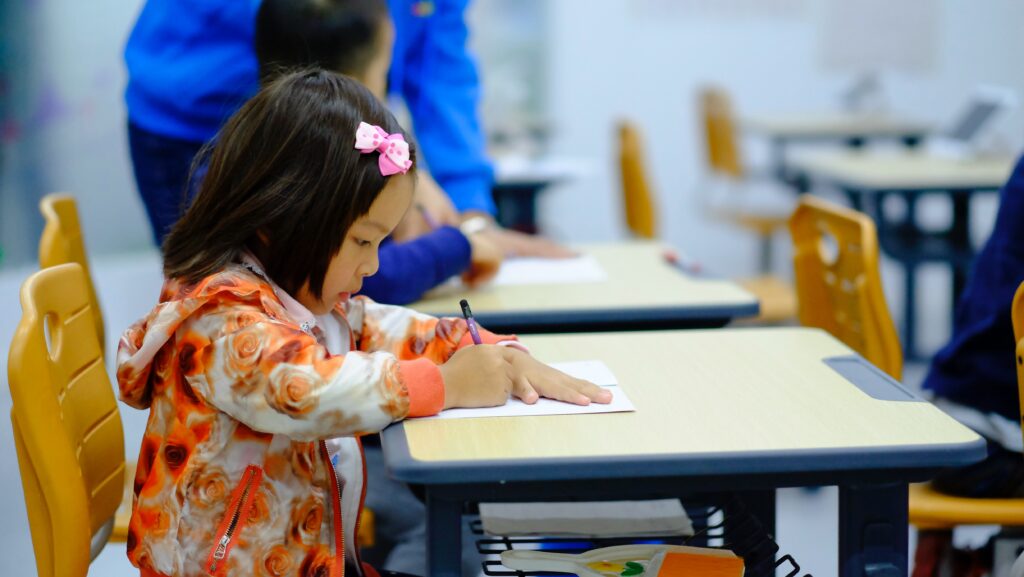 This is an image of a young girl writing in a classroom while a teacher walks around looking at student work from the blog post What is Writing Workshop? for Ms. Dorismond's Virtual Corner.